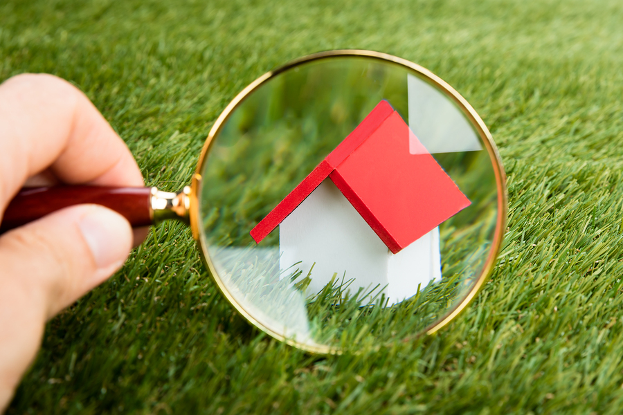 Person Hands With Magnifying Glass Inspecting A Model House On Grassy Field